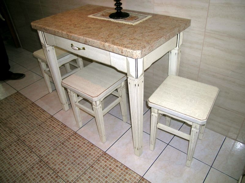 Stół kuchenny z blatem z jasnego granitu i trzema drewnianymi stołkami w stylu shabby chic, widok z boku, na blacie dekoracja.