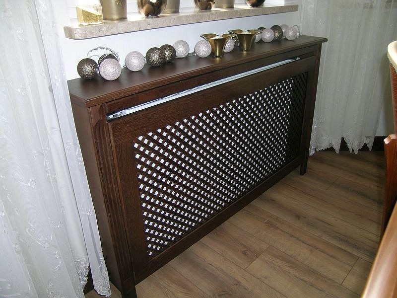 Maskownica grzejnika z ciemnego drewna z ażurowym frontem, stojąca na drewnianej podłodze, z dekoracjami na górze i zasłoną w tle.