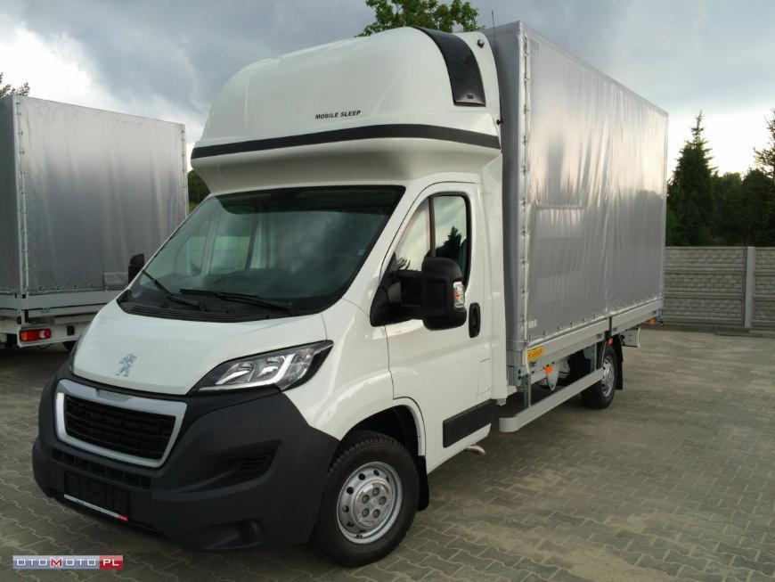 Biały bus Peugeot Boxer z zabudową plandekową i kabiną sypialną Mobile Sleep, zaparkowany na kostce brukowej, z innym busem w tle.