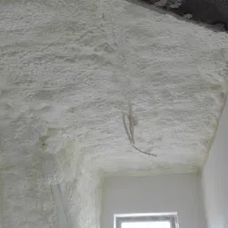 Świeżo nałożona, jasna izolacja na poddaszu, widoczne przewody elektryczne zwisające z sufitu oraz fragment okna w dolnej części kadru.