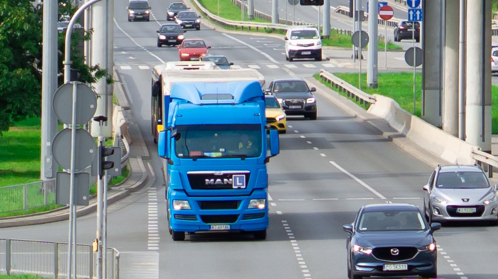 Niebieska ciężarówka MAN z literą 'L' na drzwiach kabiny, jadąca po trzypasmowej jezdni w ruchu miejskim, widziana z góry.