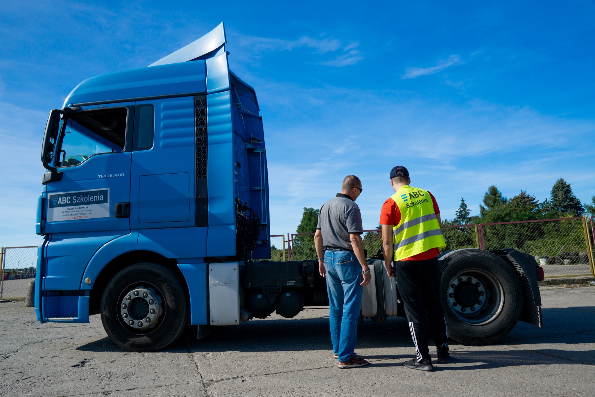 Niebieski ciągnik siodłowy MAN TGX 18.400 z logo firmy szkoleniowej ABC Szkolenia, obok którego stoją dwie osoby, jedna w kamizelce odblaskowej z napisem ABC Szkolenia, na tle błękitnego nieba...