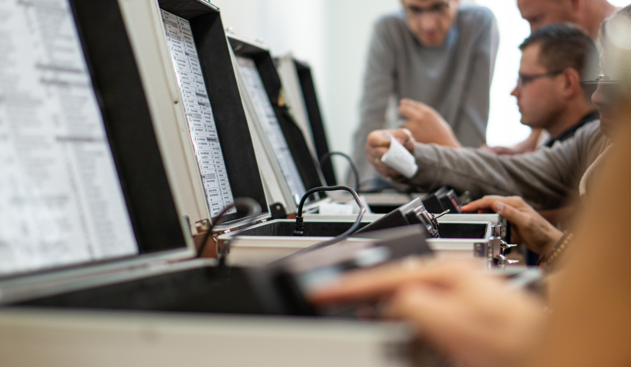 Uczestnicy kursu zawodowego przy stanowiskach egzaminacyjnych z otwartymi walizkami, zawierającymi dokumentację i przewody, podczas praktycznego egzaminu.