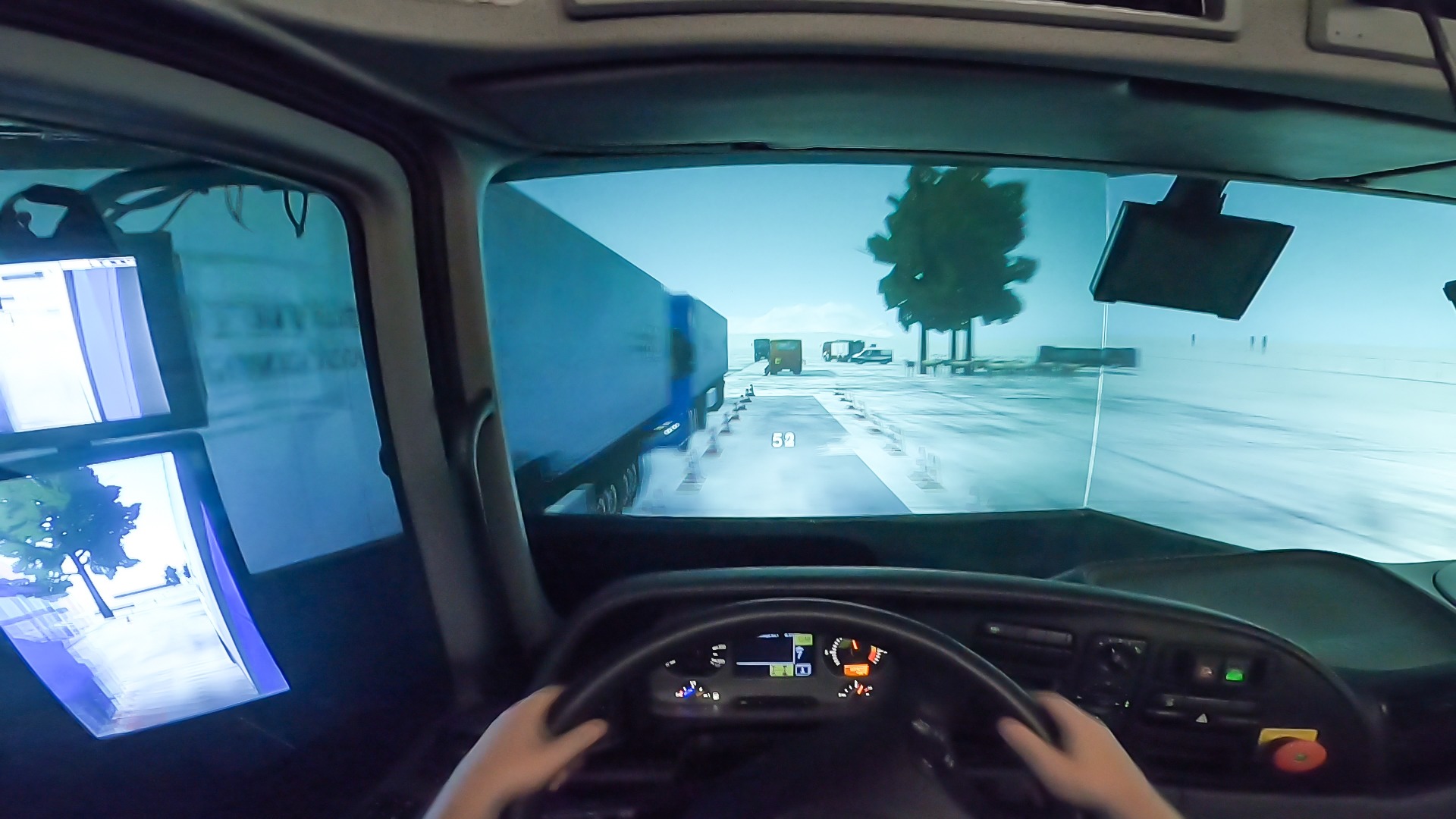 Widok z kabiny symulatora jazdy ciężarówką, z widocznym kokpitem, kierownicą i ekranami wyświetlającymi wirtualną trasę z innymi pojazdami i elementami otoczenia.