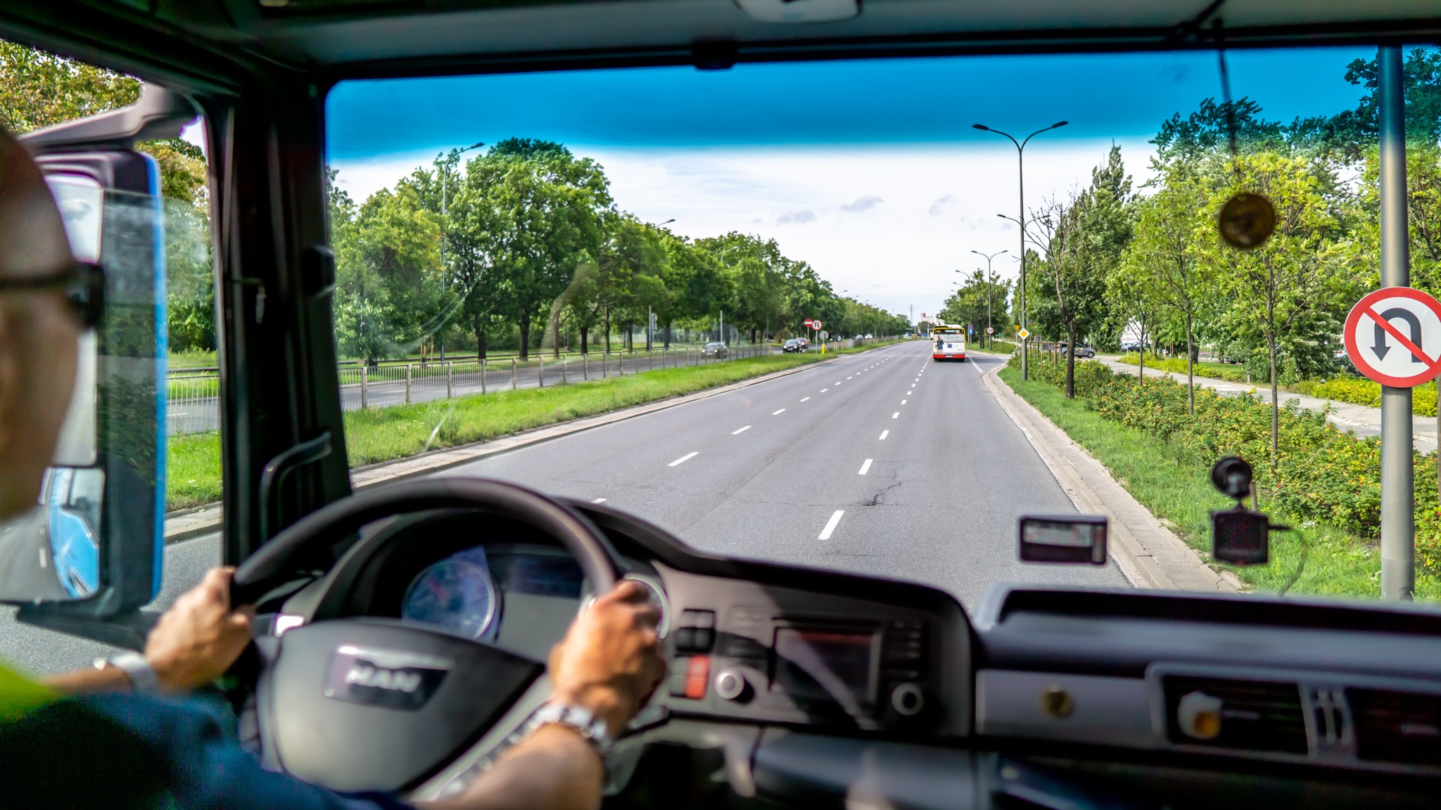 Widok z kabiny autobusu na prostą drogę z drzewami i znakiem zakazu zawracania, w oddali widoczny inny autobus.