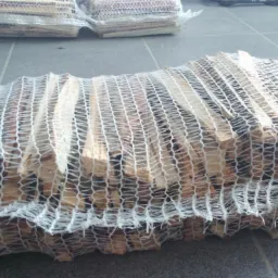 Worki z pociętym drewnem kominkowym, ułożone na szarym podłożu. Widoczne kawałki drewna przez białą siatkę worka. U góry kadru kolejne worki z drewnem.