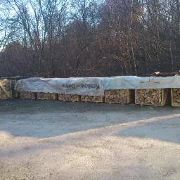 Ułożone drewno kominkowe w drewnianych skrzyniach, częściowo przykryte plandeką z nadrukiem, na tle drzew w słoneczny dzień.