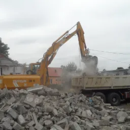 Żółta koparka Zeppelin ZM 15 ładuje gruz do ciężarówki na tle częściowo zburzonego budynku i pochmurnego nieba. Widoczny stos gruzu i sąsiednie domy.