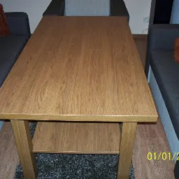 Stolik kawowy z blatem imitującym drewno, ustawiony między dwoma szarymi sofami, widoczne nogi w kolorze blatu i dolna półka, na lewo fragment osoby w różowo-niebieskich skarpetkach.