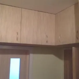 Jasne, drewniane szafki kuchenne podwieszane, z uchwytami w kształcie łuków, zamontowane nad drzwiami z oświetleniem w górnej części.