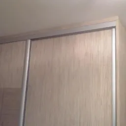 Jasna szafa przesuwna na wymiar z drewnopodobnymi frontami, srebrne prowadnice, półka z ubraniami z boku.