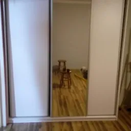 Biała szafa wnękowa z przesuwnymi drzwiami, jedno skrzydło lustrzane, widoczne dwa taborety w odbiciu, parkiet na podłodze.