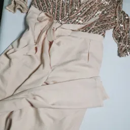 Beżowy kombinezon damski z błyszczącą górą w cekiny, wiązany w pasie, leżący na białym tle.