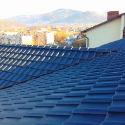 Niebieski dach z blachodach&oacute;wki w promieniach słońca, w tle zabudowa miejska i g&oacute;ry. Widoczne detale łączeń i faktura blachy. Realizacja dekarska.