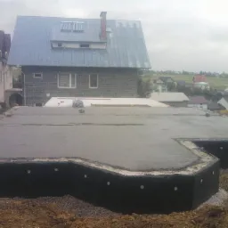 Zalany fundament pod dom z hydroizolacją w czarnej folii z widocznymi mocowaniami. W tle budynek w trakcie budowy i zabudowa jednorodzinna na wzgórzu.