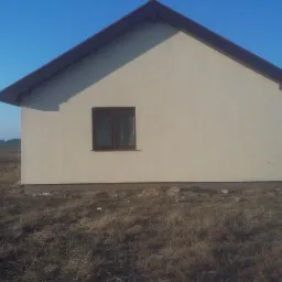 Boczny widok domu z beżową elewacją i brązowym dachem w słoneczny dzień. Widoczne okno z brązową ramą i trawa na pierwszym planie. W tle błękitne niebo i sąsiednie budynki.