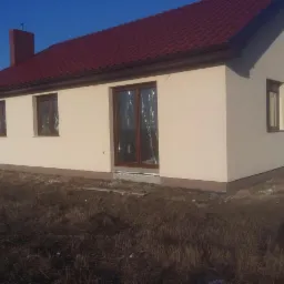 Beżowy dom jednorodzinny z brązowymi oknami i dachem z czerwonej dachówki, widok z zewnątrz. Okno zabezpieczone folią. Widoczne wykończenie elewacji.