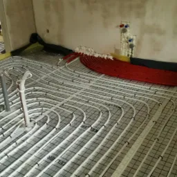 Układanie białych rur ogrzewania podłogowego na czarnej siatce z żółtą folią brzegową, czerwone rury w tle, pianka montażowa przy ścianie, surowy stan wykończenia wnętrza.