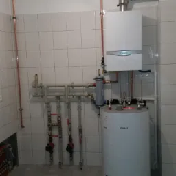 Biały, wiszący piec gazowy marki Vaillant z podłączonym kominem i zbiornikiem na wodę, instalacja z rur miedzianych i zaworów, szare płytki na ścianach i podłodze w kotłowni.