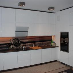 Meble Zyubra - Biała kuchnia na wymiar z błyszczącymi frontami, ciemnym blatem i fototapetą z motywem retro, oświetlona halogenami. Widoczny piekarnik w zabudowie.