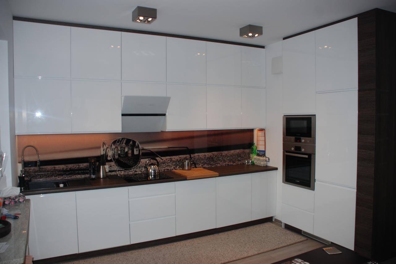 Biała kuchnia na wymiar z błyszczącymi frontami, ciemnym blatem i fototapetą z motywem retro, oświetlona halogenami. Widoczny piekarnik w zabudowie.