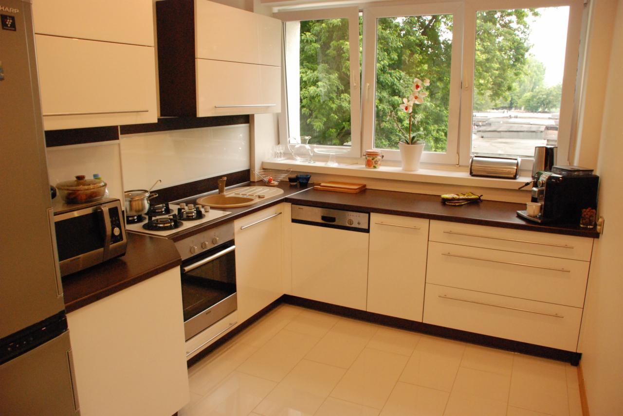 Jasna kuchnia na wymiar z ciemnym blatem i sprzętami AGD. Widok na okno z kwiatem, ekspres do kawy i toster. Ujęcie z perspektywy narożnika.