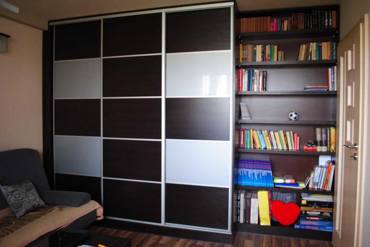 Wnętrze pokoju z dużą szafą wnękową z przesuwnymi drzwiami w kolorze ciemnego drewna i szarego szkła, obok regał z książkami i dekoracjami.