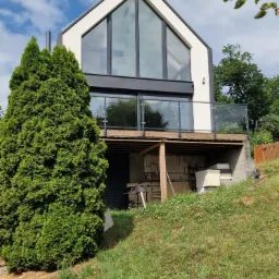 Nowoczesny dom z dużymi przeszkleniami, widok z dołu na elewację z balkonem z szklaną balustradą i wysokimi oknami w szczycie, na pierwszym planie trawnik i tuja.