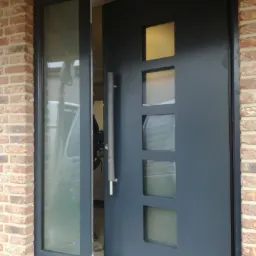 Grafitowe drzwi wejściowe z czterema kwadratowymi przeszkleniami, zamontowane w ścianie z cegły, widoczny fragment montażu i odbicie samochodu w szybie.