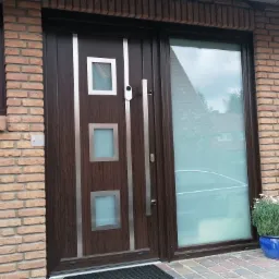 Brązowe drzwi wejściowe z trzema kwadratowymi przeszkleniami i długim uchwytem, zamontowane w ścianie z cegły klinkierowej, obok okno z brązową ramą i skrzynka na listy ze stali nierdzewnej.