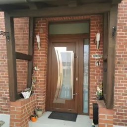 Brązowe drzwi wejściowe z dekoracyjnym, złotym elementem, zamontowane w ceglanym budynku z drewnianym zadaszeniem i ozdobnymi detalami.