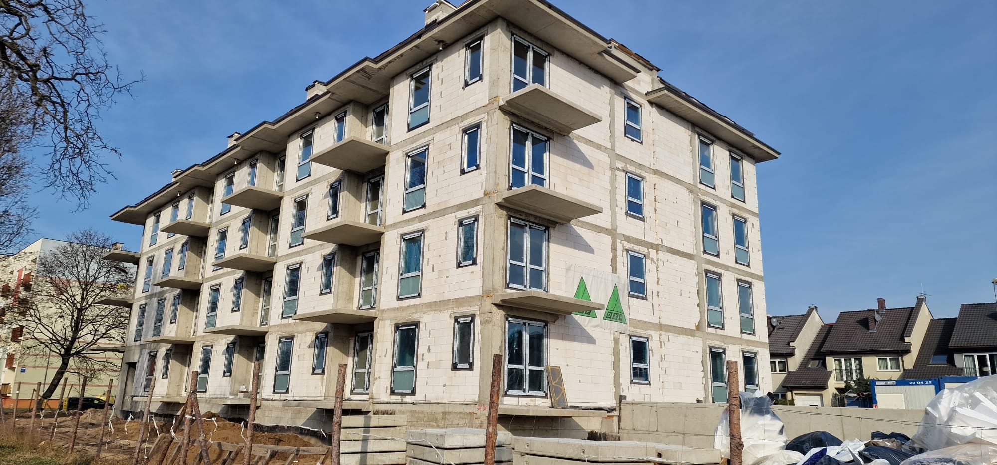 Wielopiętrowy budynek w trakcie budowy z zamontowanymi oknami, widoczne balkony i materiały budowlane na pierwszym planie.