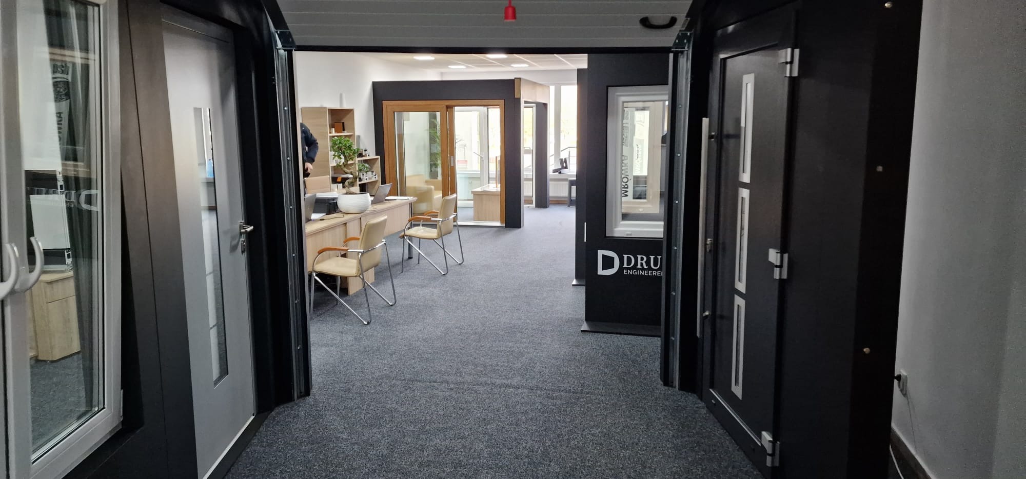 Wnętrze salonu z ekspozycją okien i drzwi w różnych stylach i kolorach, widok przez otwarte drzwi na biurko z bonsai i krzesłami.