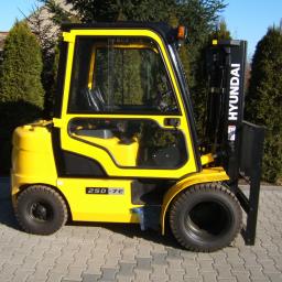 Jg Serwis-Truck - Żółty wózek widłowy Hyundai 25D-7E na kostce brukowej, z zielonymi krzewami w tle. Widok z boku, oświetlenie dzienne.