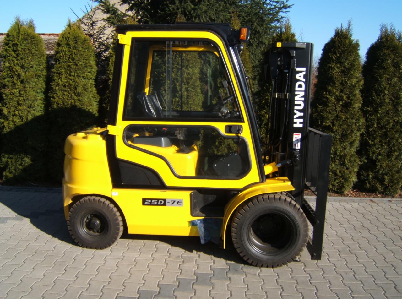 Żółty wózek widłowy Hyundai 25D-7E na kostce brukowej, z zielonymi krzewami w tle. Widok z boku, oświetlenie dzienne.