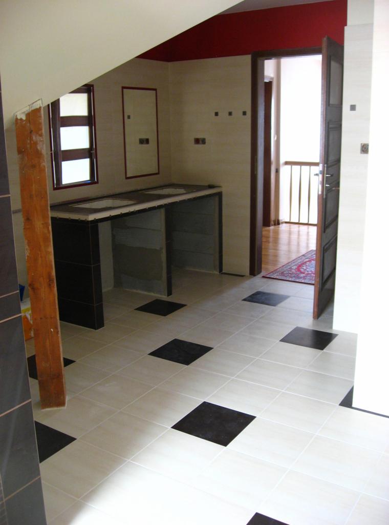 Łazienka w trakcie wykańczania: jasne płytki z czarnymi akcentami na podłodze, zabudowa pod umywalkę w czerni, widoczna konstrukcja, otwarte drzwi do innego pomieszczenia.