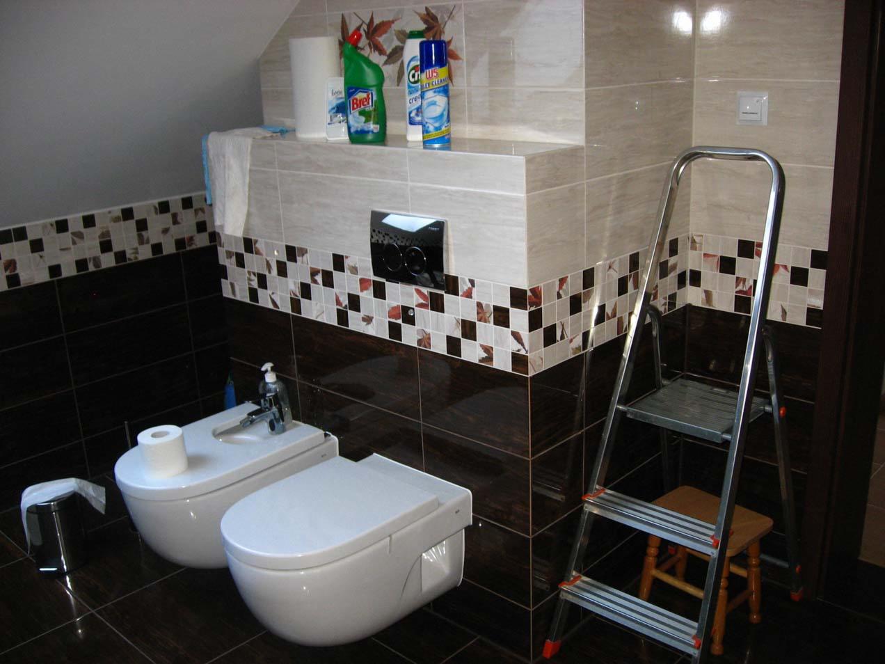 Wykończona łazienka z mozaiką, brązowymi płytkami i białymi sanitariatami. Na półce środki czystości i drabina oparta o ścianę. Widok na wnętrze łazienki.