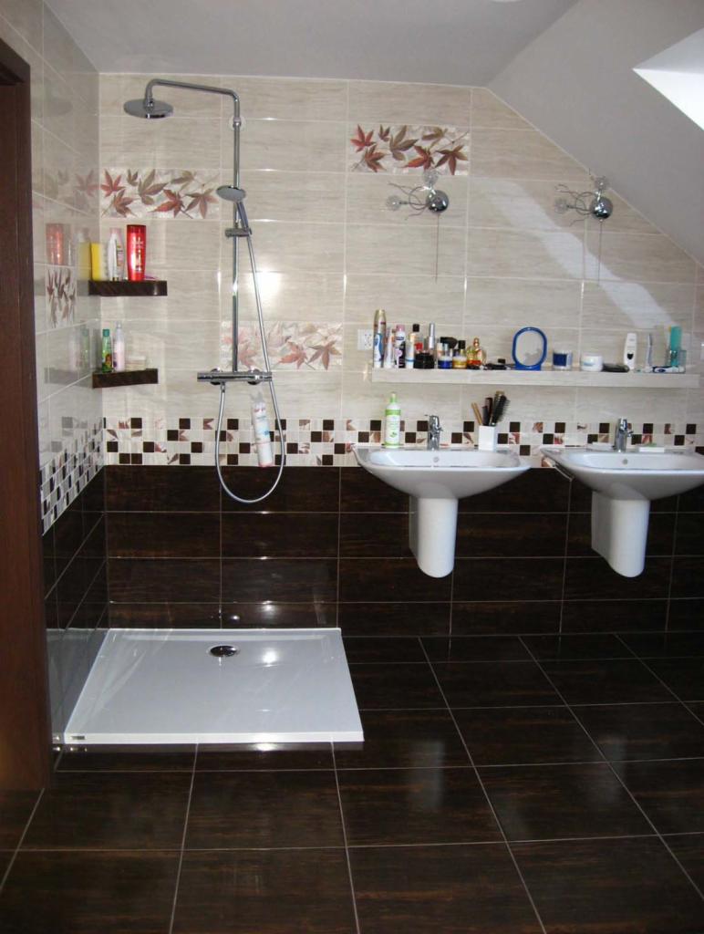 Ukończona łazienka z brązowymi płytkami, białą brodzikiem i dwoma umywalkami. Na ścianach beżowe płytki z motywem liści i półki z kosmetykami.