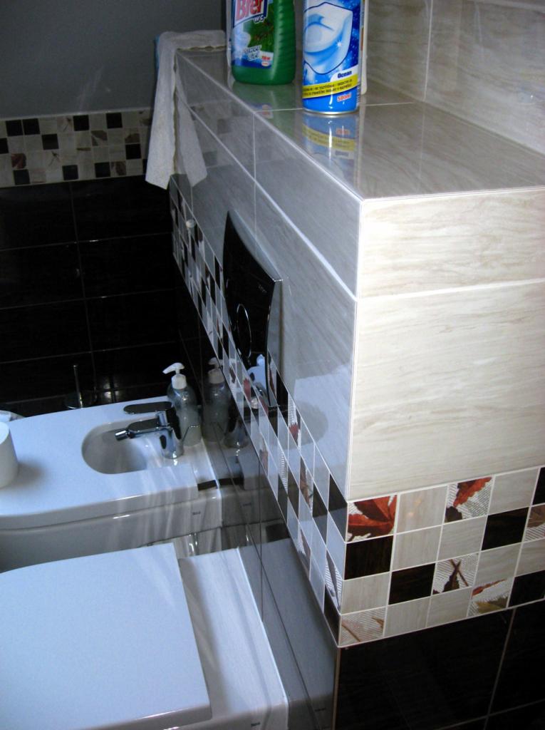 Wykończona łazienka: bidet, toaleta i umywalka z blatem z płytek imitujących drewno. Na blacie środki czystości. Dekoracyjne płytki w szachownicę.