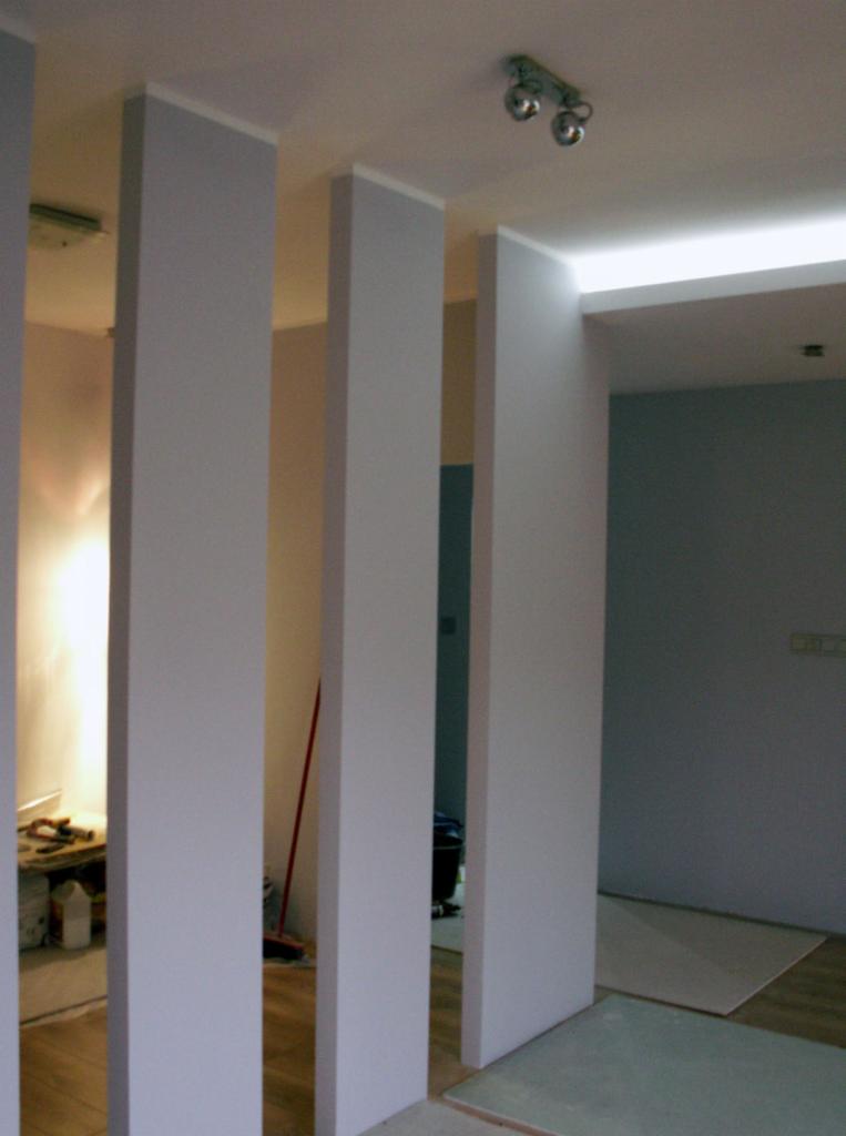 Wnętrze z trzema kolumnami w odcieniach szarości, oddzielającymi pomieszczenia. Widoczne oświetlenie sufitowe i fragment podłogi w trakcie prac wykończeniowych.