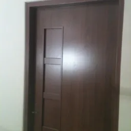 Ciemnobrązowe drzwi wewnętrzne z prostokątnym, wpuszczonym panelem dekoracyjnym i srebrną klamką, osadzone w dopasowanej ościeżnicy na tle jasnej ściany.