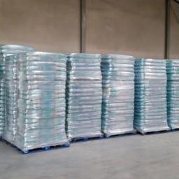 Roem - Magazyn z paletami środków czystości w przezroczystych opakowaniach foliowych, ustawionych w rzędach na niebieskich paletach. Widoczne metalowe ściany magazynu.