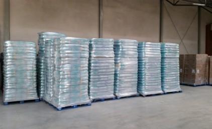 Magazyn z paletami środków czystości w przezroczystych opakowaniach foliowych, ustawionych w rzędach na niebieskich paletach. Widoczne metalowe ściany magazynu.