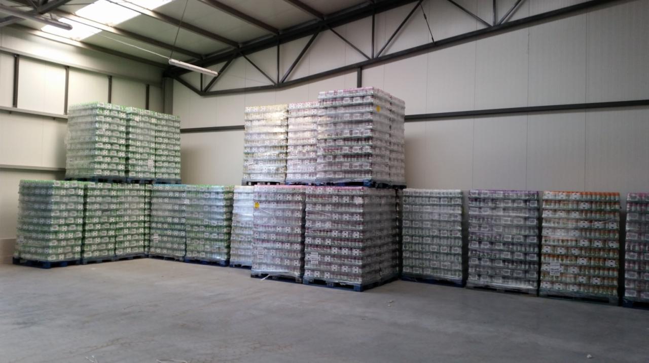 Magazyn z paletami środków czystości w folii, różne kolory, równo ułożone. Hala z metalową konstrukcją stropu i betonową posadzką.