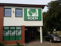 Elewacja budynku z zielonym szyldem 'ROEM' i oknami z powtarzającym się logo, szara kostka brukowa, zaparkowany samochód, drzewa w tle.