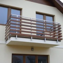 Balkon z drewniano-metalową balustradą, widok z dołu. Drewniane elementy w ciepłym odcieniu brązu, metalowe słupki w kolorze srebrnym. Jasna elewacja budynku w tle.