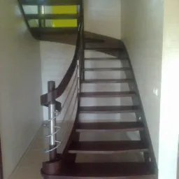 Kręte schody z ciemnego drewna z metalową balustradą, widoczne z dołu, z żółtym akcentem w tle. Minimalistyczny design.