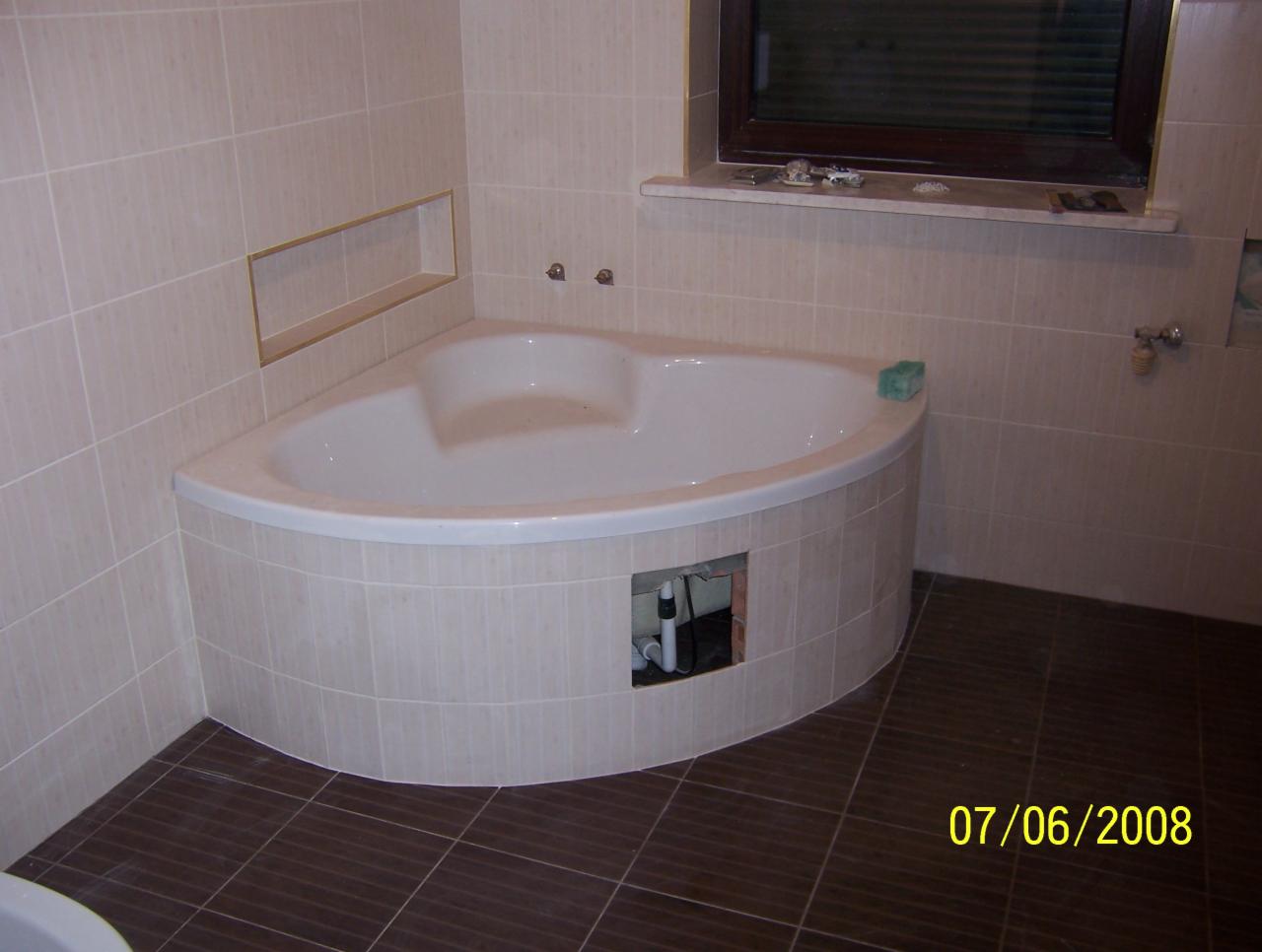 Narożna wanna obudowana płytkami w łazience z brązową podłogą i beżowymi ścianami, widoczne instalacje pod wanną przez otwór rewizyjny.