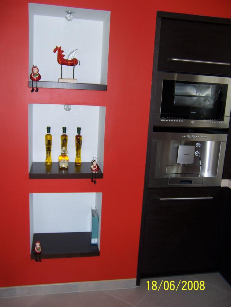 Czerwona ściana z trzema wnękami, w których umieszczono dekoracje: figurkę konia ze skrzydłami, butelki z oliwą i małą figurkę, obok zabudowa kuchenna z piekarnikiem i ekspresem do kawy.