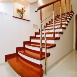 Klatka schodowa z drewnianymi stopniami w ciepłym odcieniu i metalową balustradą, oświetlona delikatnym światłem. Na ścianie dekoracja kwiatowa.